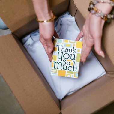 A person holds a thank you card above items in an open shipping box, suggesting care in sending.