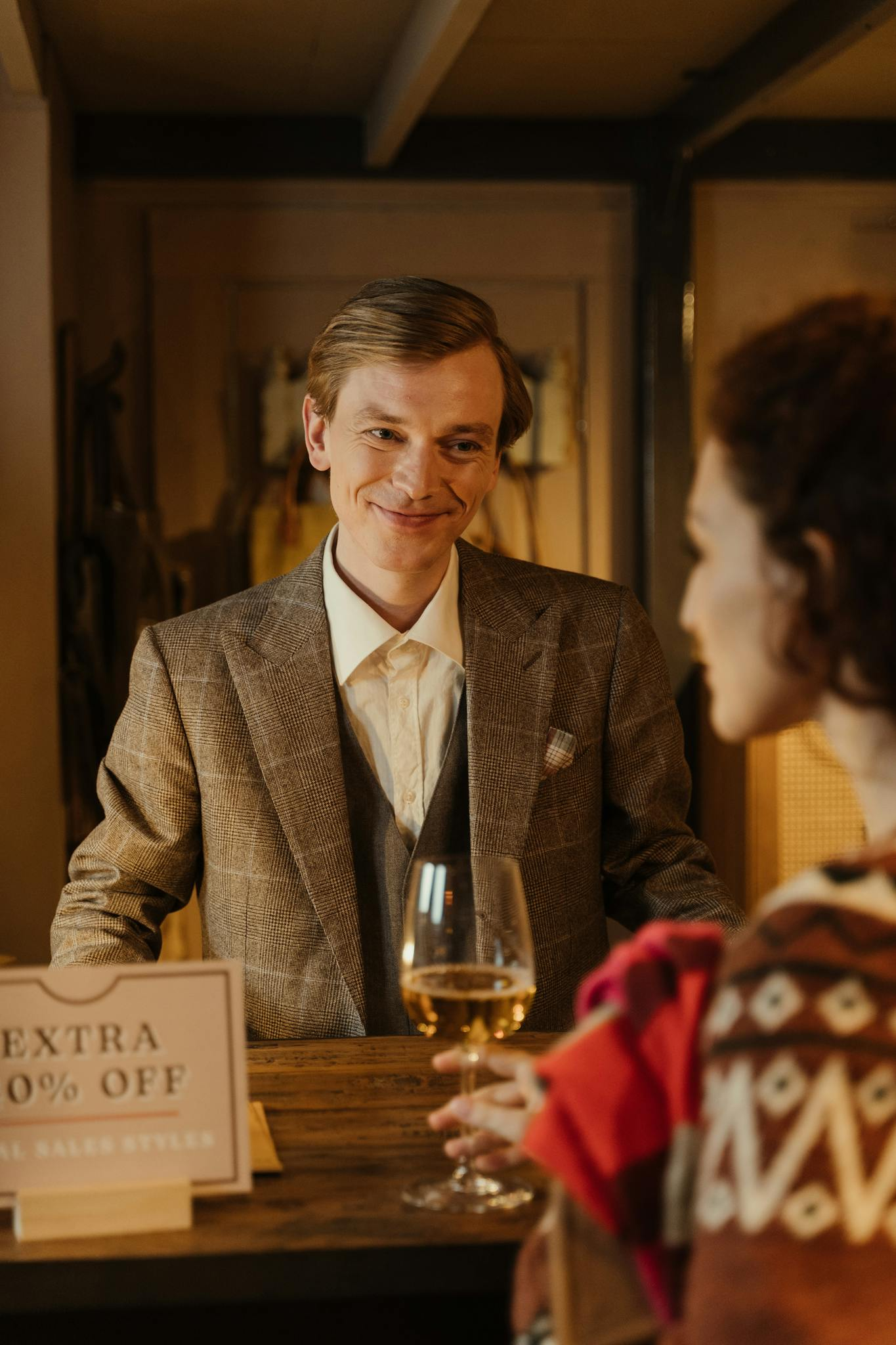 A smiling salesman offers a wine glass to a customer at a shop counter.